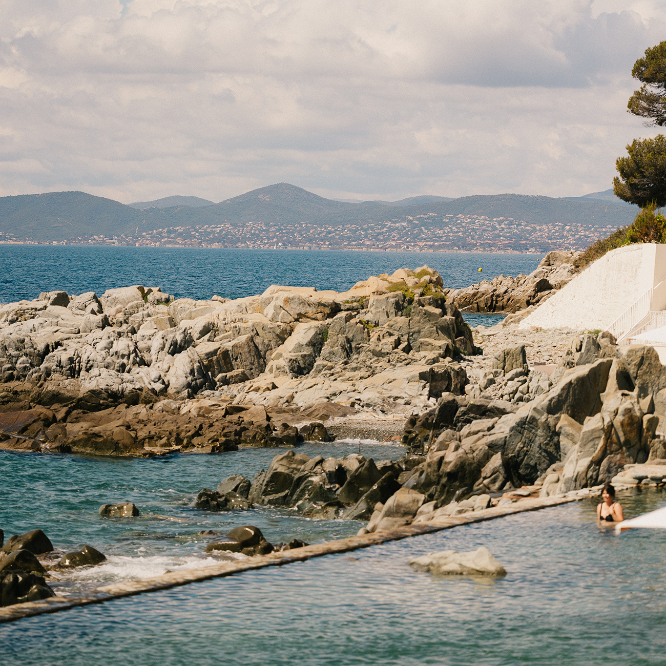 Rocky coastline with turquoise sea, distant mountains, and a cloudy sky. A person relaxes on a paddleboard near the shore.
