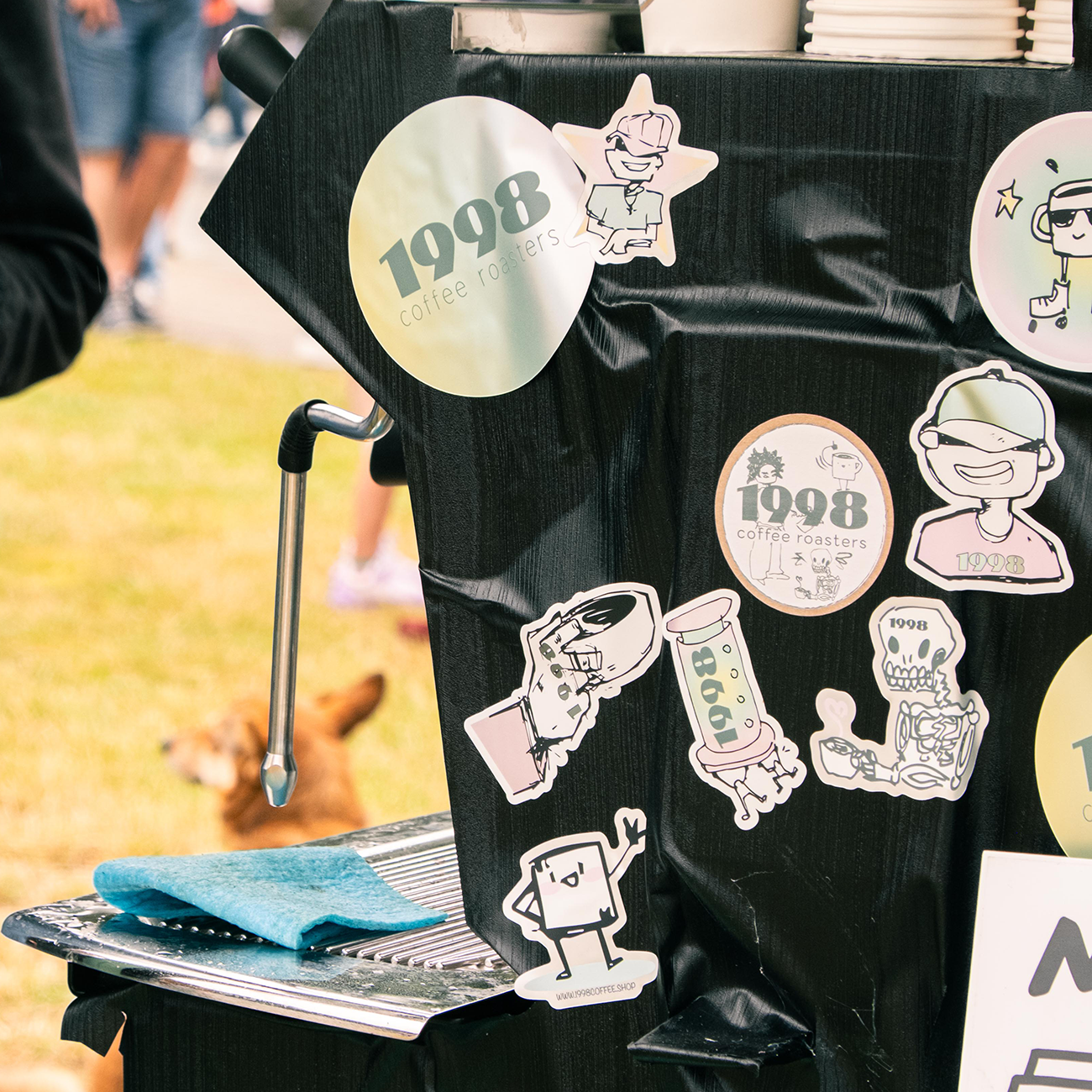Coffee cart decorated with playful die cut stickers, including "1998 coffee roasters," cartoon characters, and a coffee cup, with a grassy background.