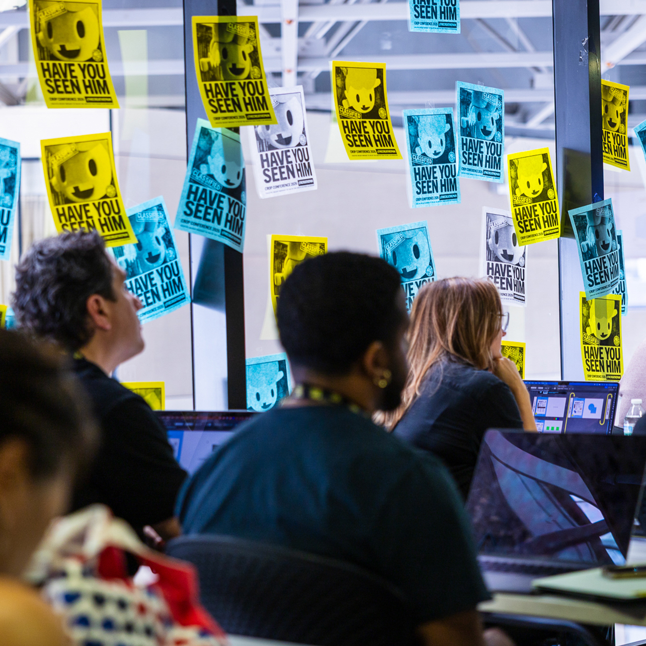 Workshop attendees at laptops in front of a glass wall covered in “Have you seen him” posters