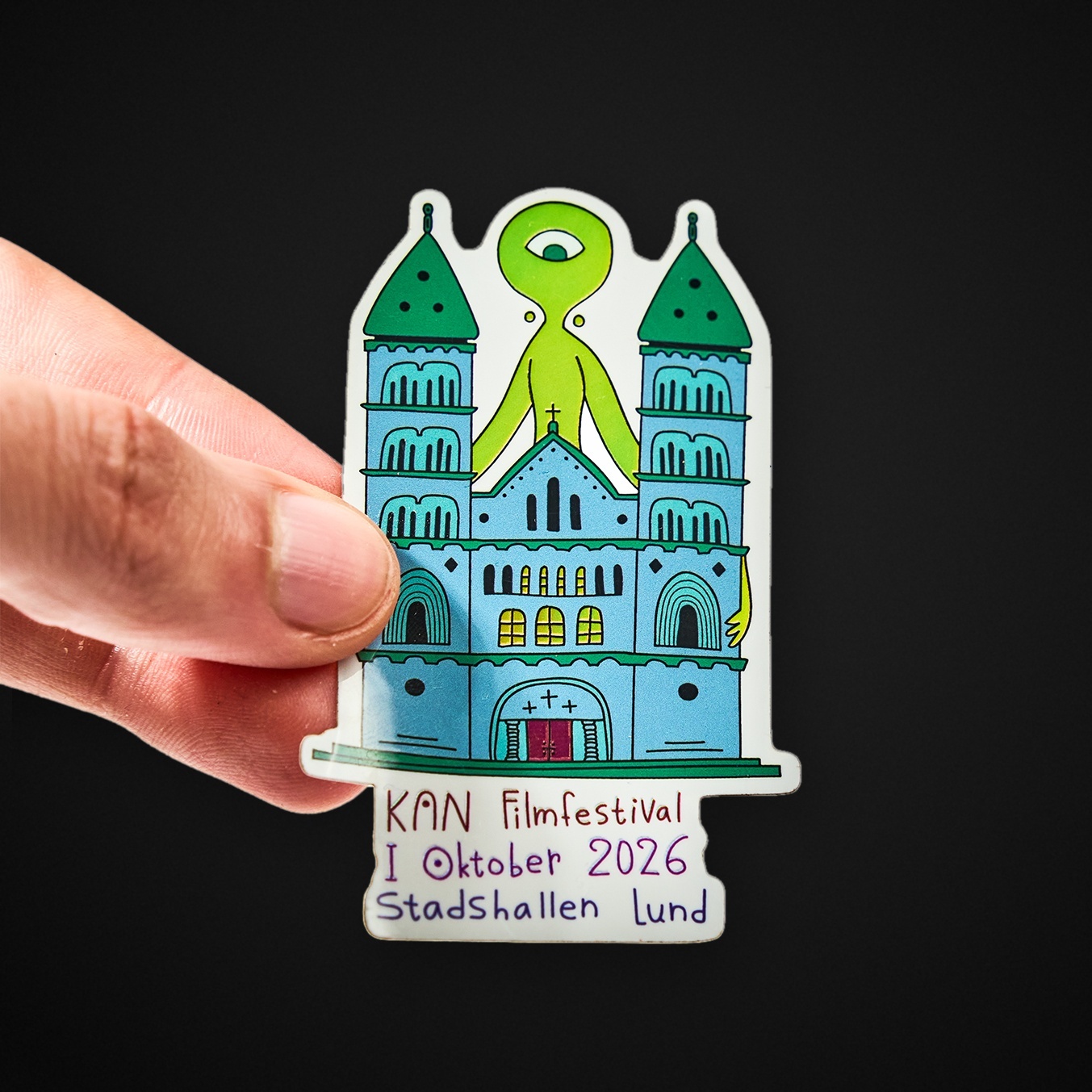 Hand holding a die-cut sticker of a cathedral with a green one-eyed creature, reading "KAN Filmfestival I Oktober 2026 Stadshallen Lund"