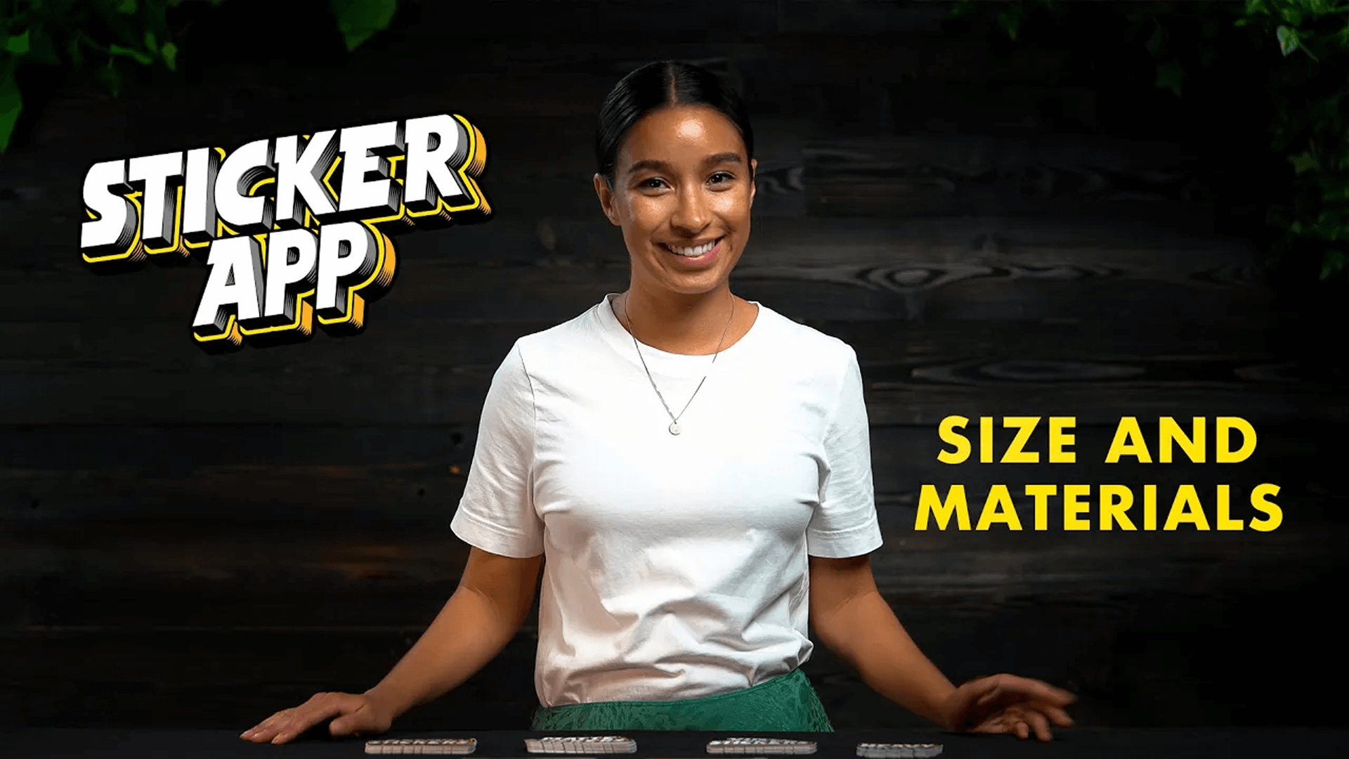 A person in a white shirt stands behind a table with stickers. Text reads "Sticker App" and "Size and Materials" on a dark wood background.